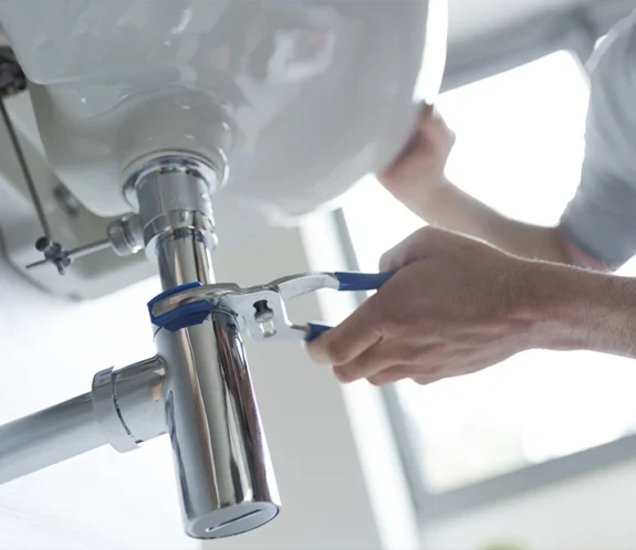 En person använder en rörtång för att laga ett vattenlås under ett handfat. Rörmokare på Gotland.