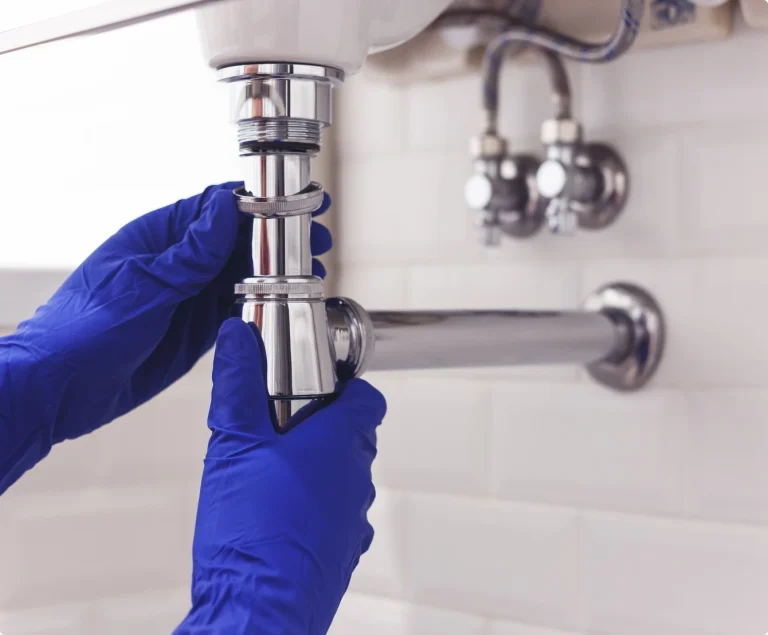 Händer i blå arbetshandskar monterar ett vattenlås i krom. Rörmokare på Gotland.