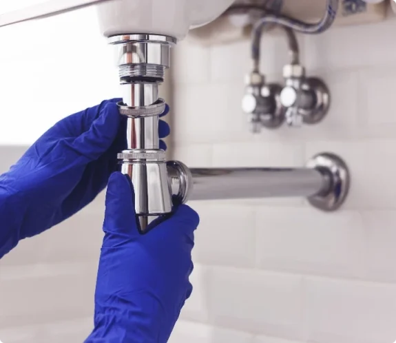 Händer i blå arbetshandskar monterar ett vattenlås i krom. Rörmokare på Gotland.